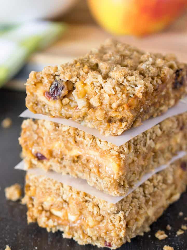 Apple crumb bars stacked three high with wax paper between them and an apple in the background.