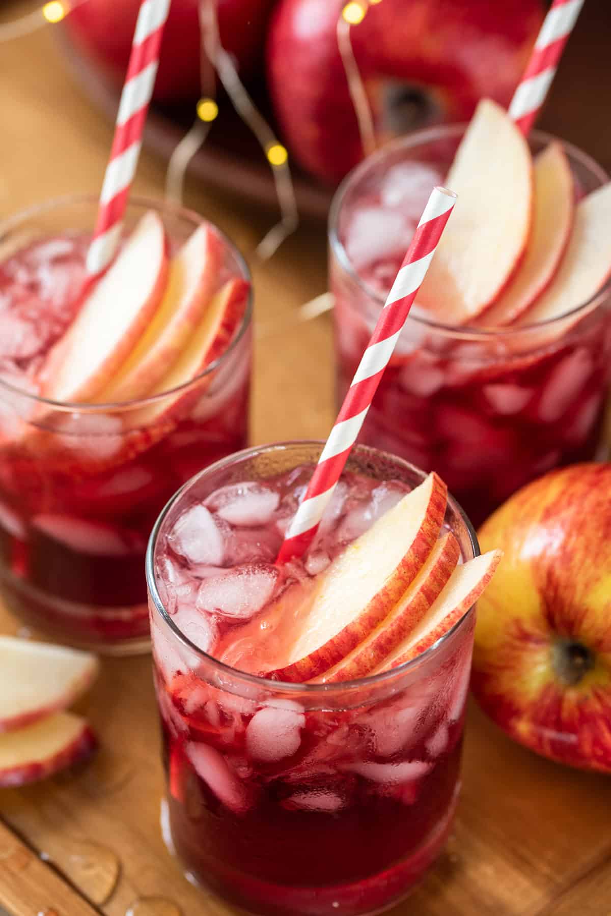 Pomegranate Crown Apple Cocktail over ice in rocks glasses, garnished with thinly sliced apples on wooden cutting board.