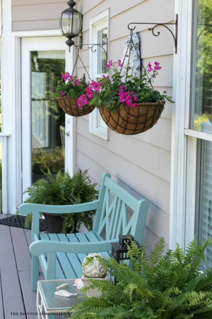 Two hanging baskets planted with seasonal pink flowers hung from wrought iron hangers on front porch of tan sided home with white trim.