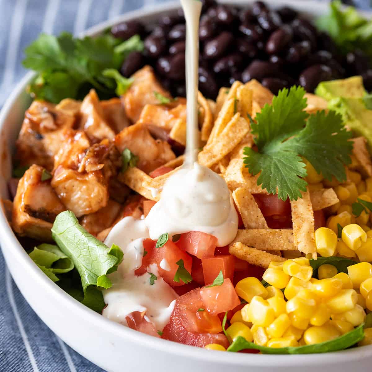 Ranch dressing being poured over a salad with corn, black beans, chicken, cilantro, and tortilla strips on it.
