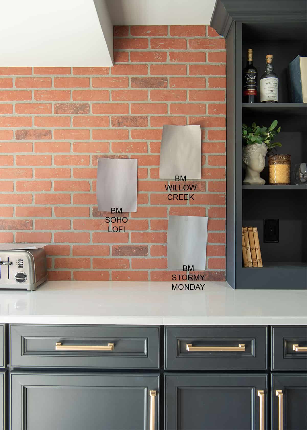Three paint samples taped against a brick wall with grey cabinets and brass handles in the background.