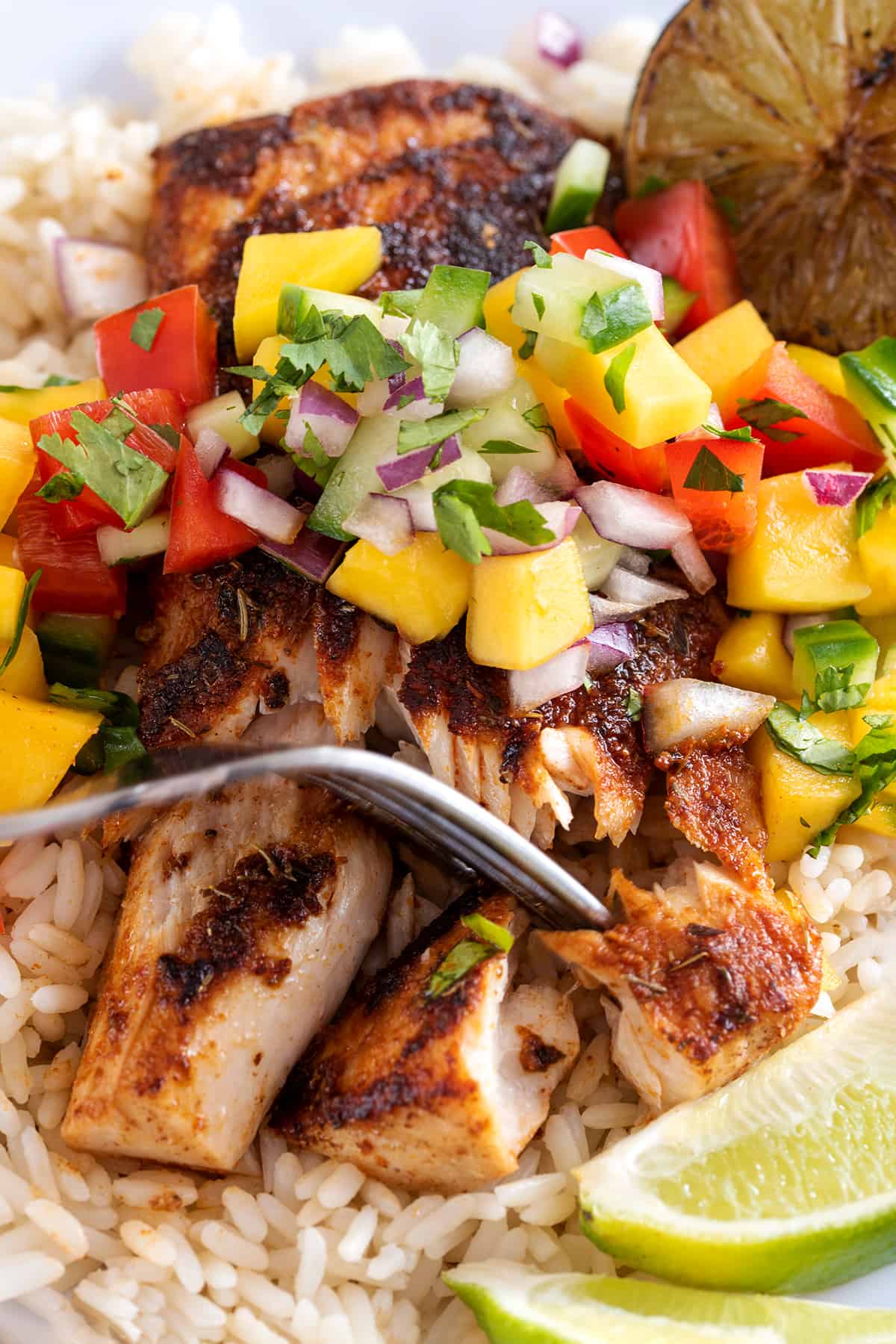 A fork pressing into a fillet of mahi mahi fish topped with cajun seasoning and mango salsa.
