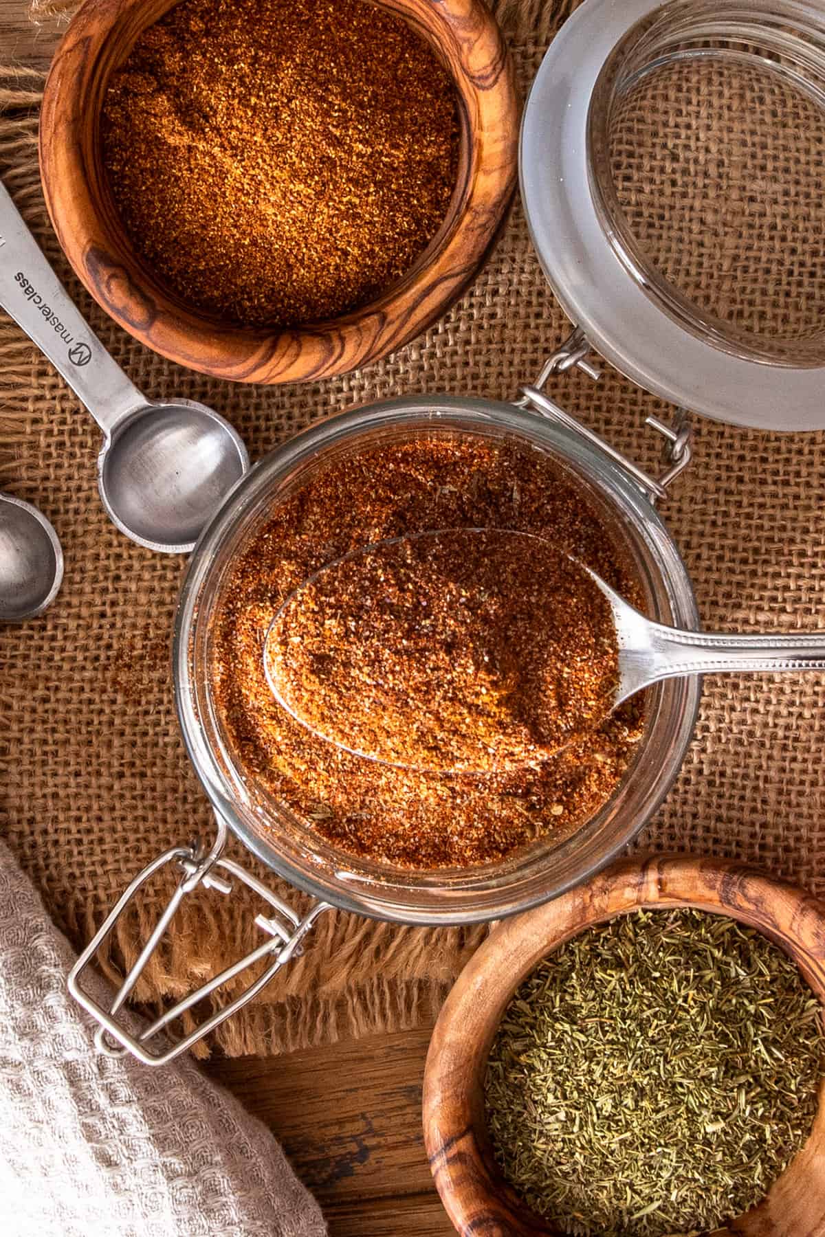 Overhead of a spoonful of cajun seasoning over top of a spice jar with bowls of herbs on each side.