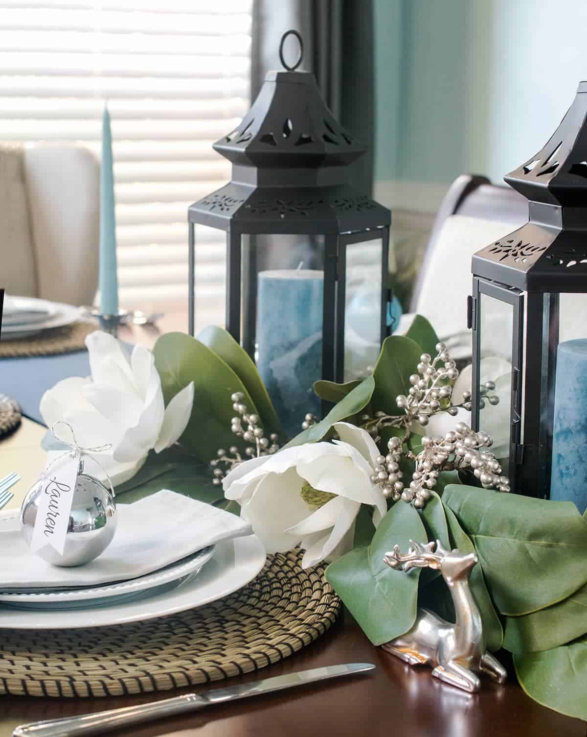 Christmas table setting with blue candles inside black lanterns. Magnolia flower garland. Silver ornament, deer, and berry accents.
