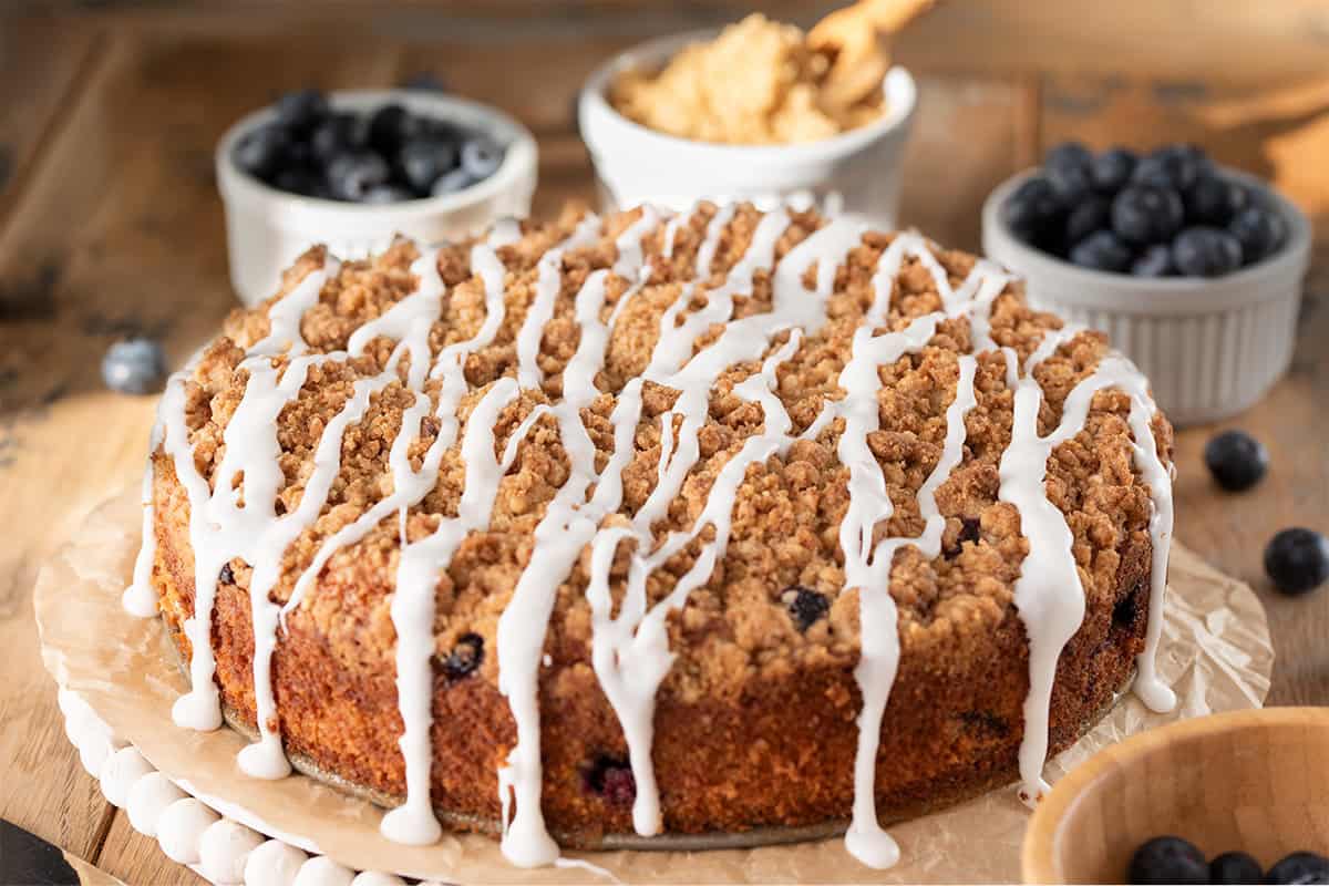 Whole blueberry sour cream coffee cake with white drizzle on top on a plate surrounded by bowls of blueberries and brown sugar.