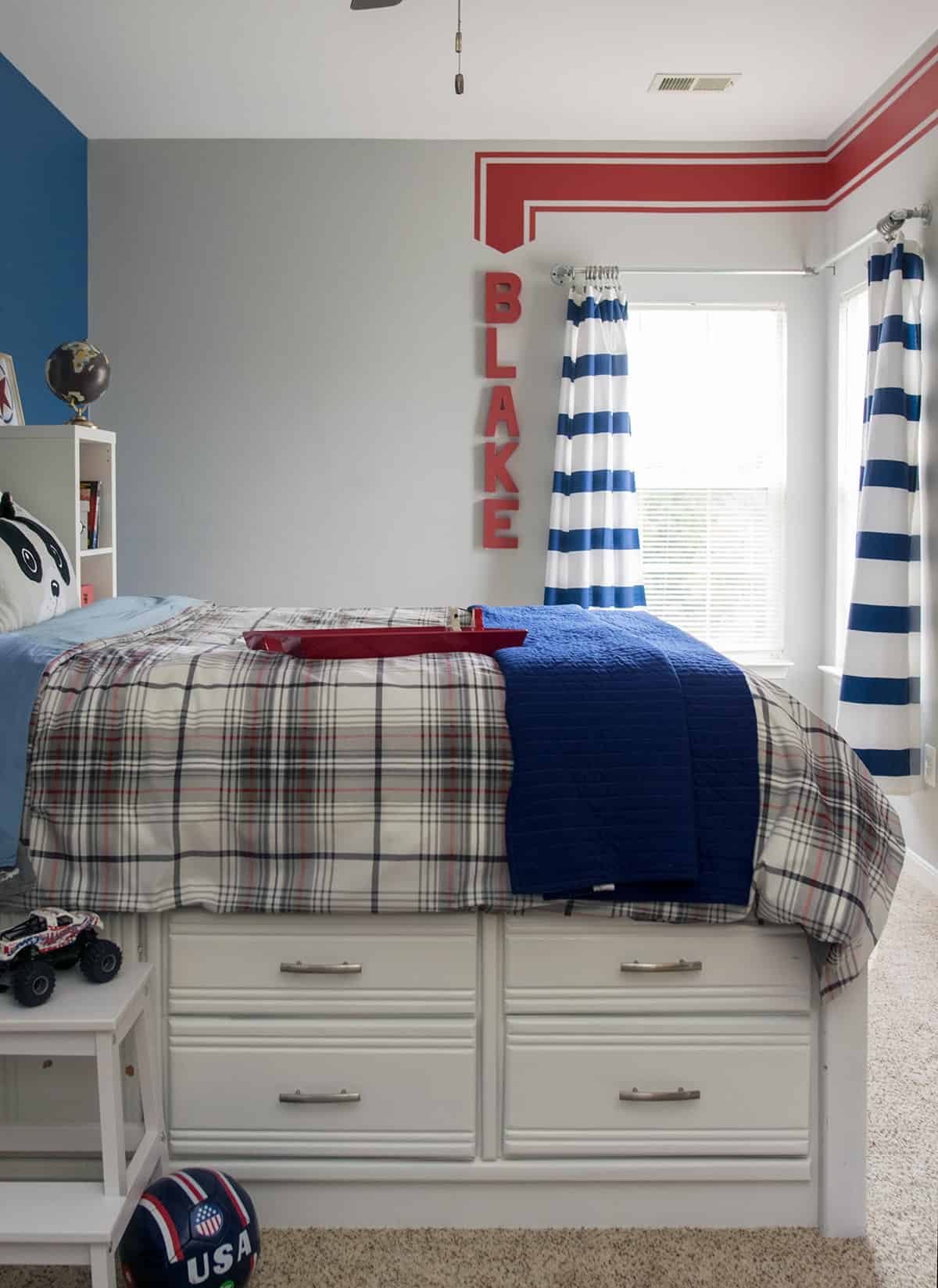 Painted white bed in little boy's room with plaid comforter and navy blanket. Dark blue and gray walls with personalized red wall stripe.