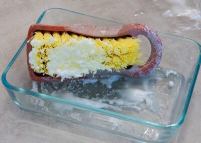 A hand sized scrub brush with baking soda solution on it for cleaning shower walls.