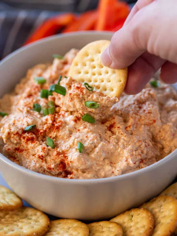 A butter cracker being dipped into cold Cajun crab dip with green onions on top.