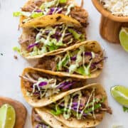 Carnitas Tacos with slaw and sauce sprinkled on top. Tacos are laid out on a white background.