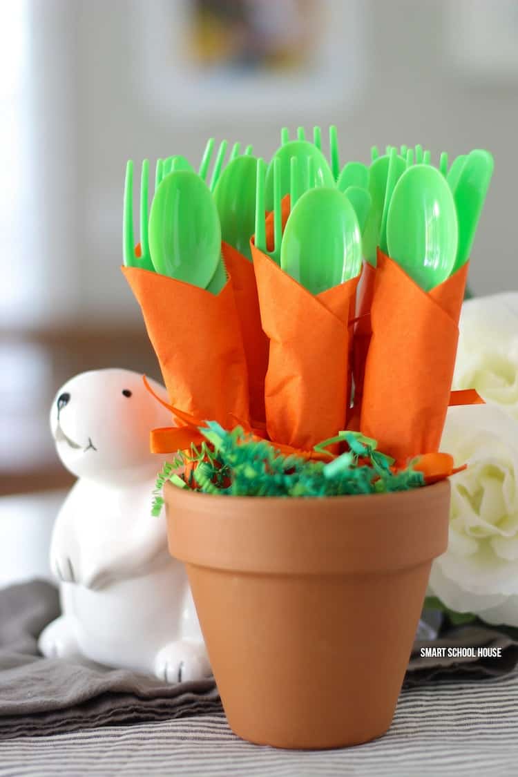 Clay pot filled with green Easter grass and green plasticware wrapped in orange napkins next to white bunny and flowers.