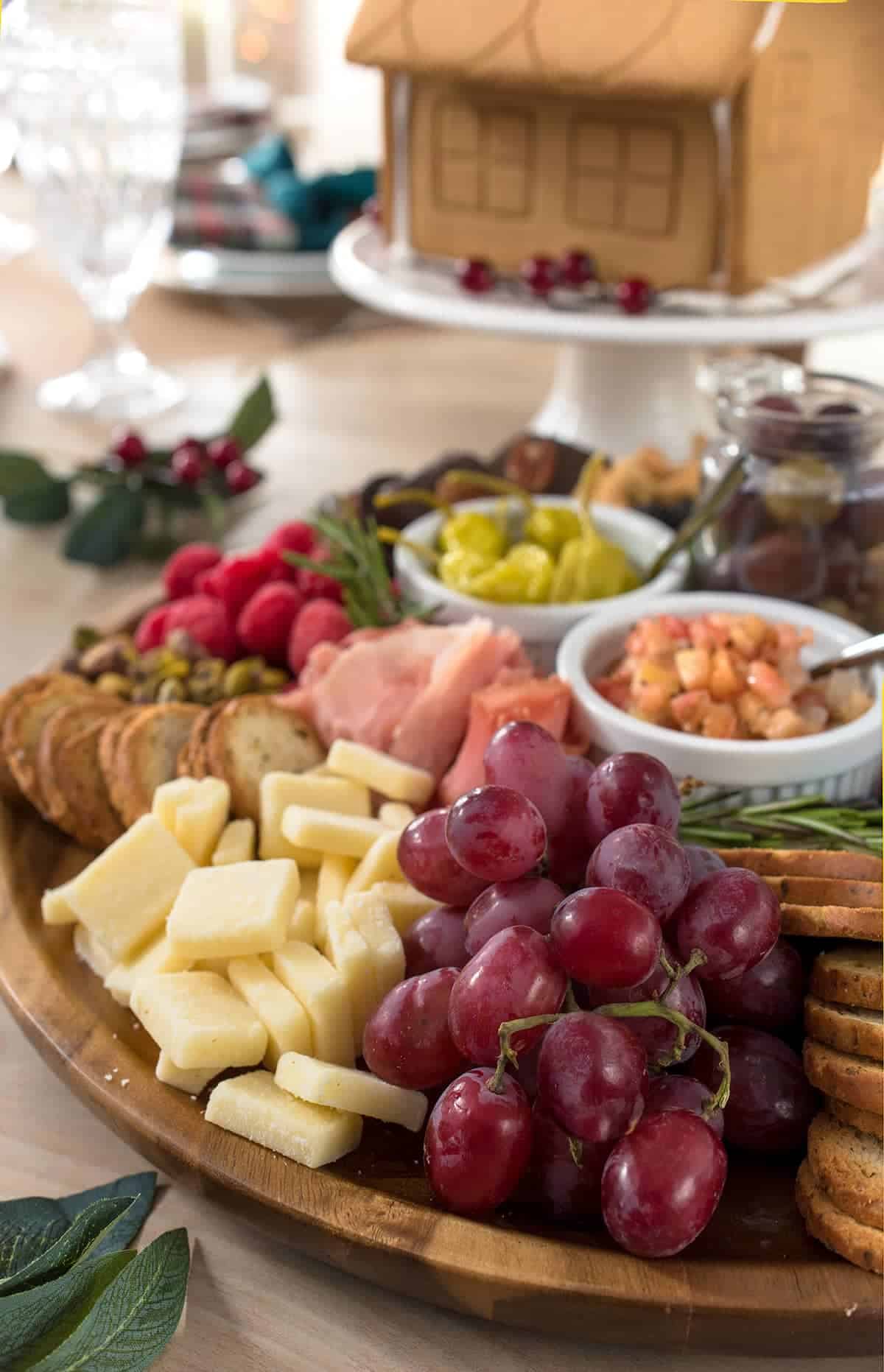 Full charcuterie board on display on dining room table. Close up of grapes, cheddar cheese, bagel crisps, pistachios and bruschetta. Crystal goblet and gingerbread house in background.