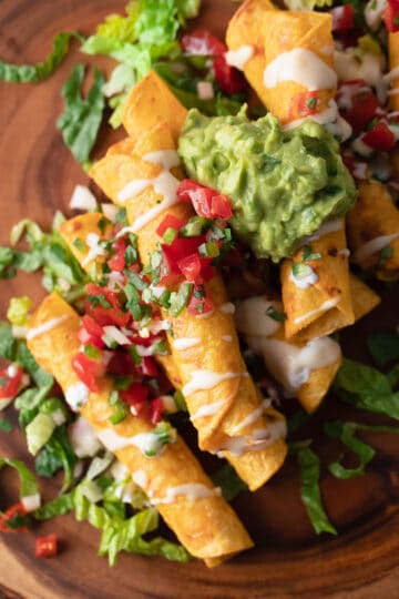 Overhead of a pile of taquitos topped with guacamole, sour cream, and diced tomatoes.