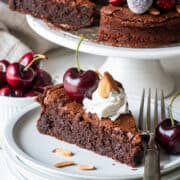 A slice of flourless chocolate almond cake with whipped cream on top in front of a full chocolate cake with cherries.