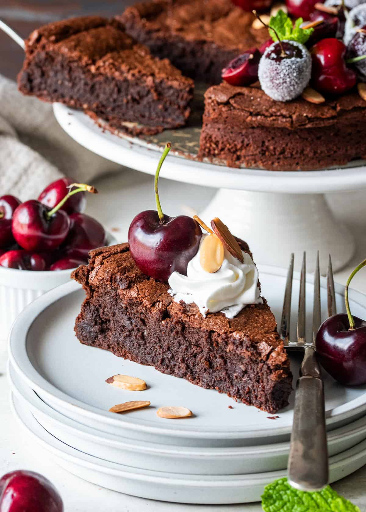 A slice of flourless chocolate almond cake with whipped cream on top in front of a full chocolate cake with cherries.