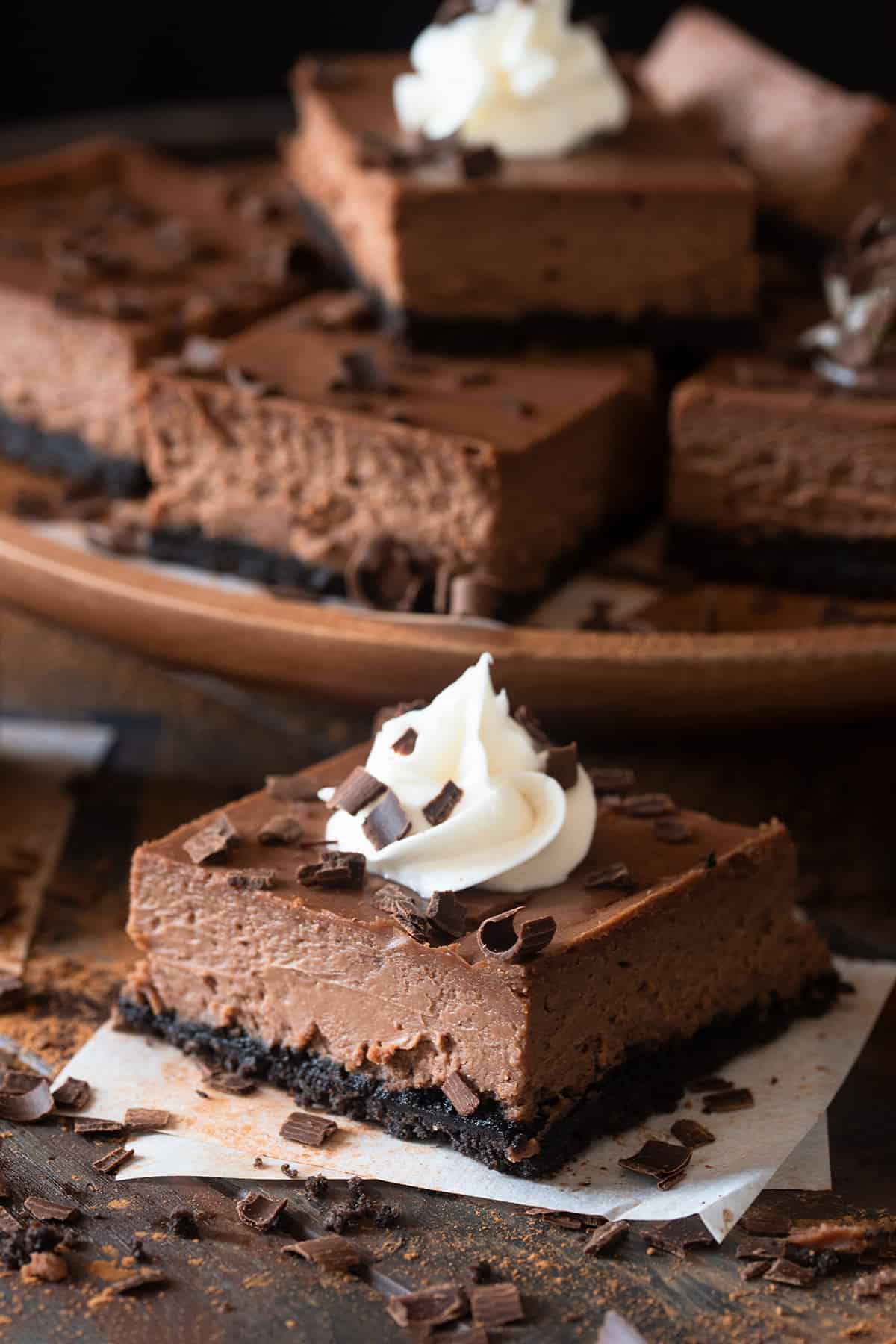 A chocolate cheesecake bar with chocolate chips sprinkled around sitting on parchment paper.