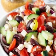Closeup of chopped greek salad with olives, tomatoes, cucumber, and feta with dressing in the background.