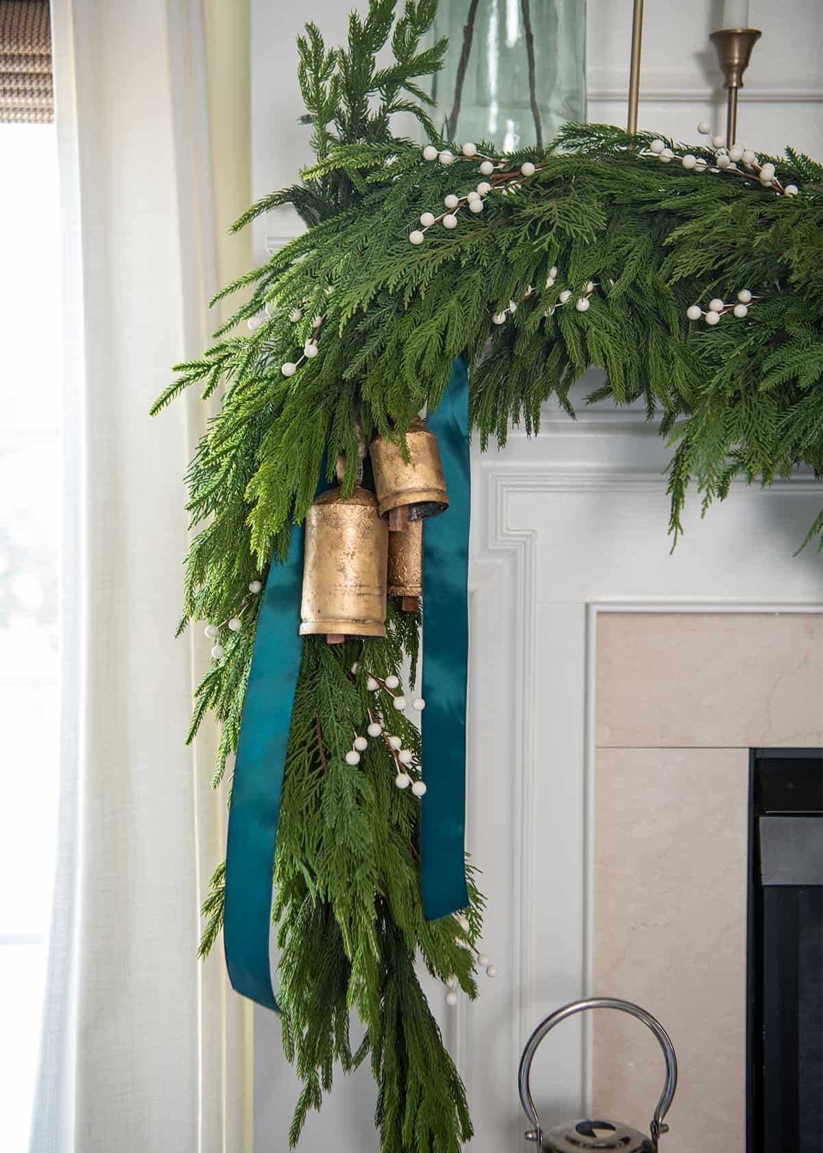 Brass antique bells and teal ribbon tucked into natural Christmas garland on a fireplace mantel.