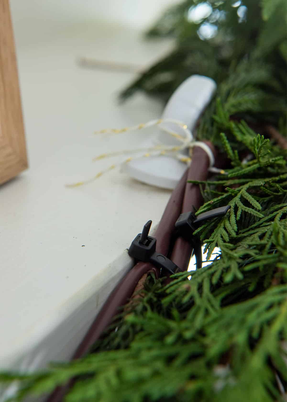 Closeup of pine greenery being secured on a mantel with zip ties and command hooks.