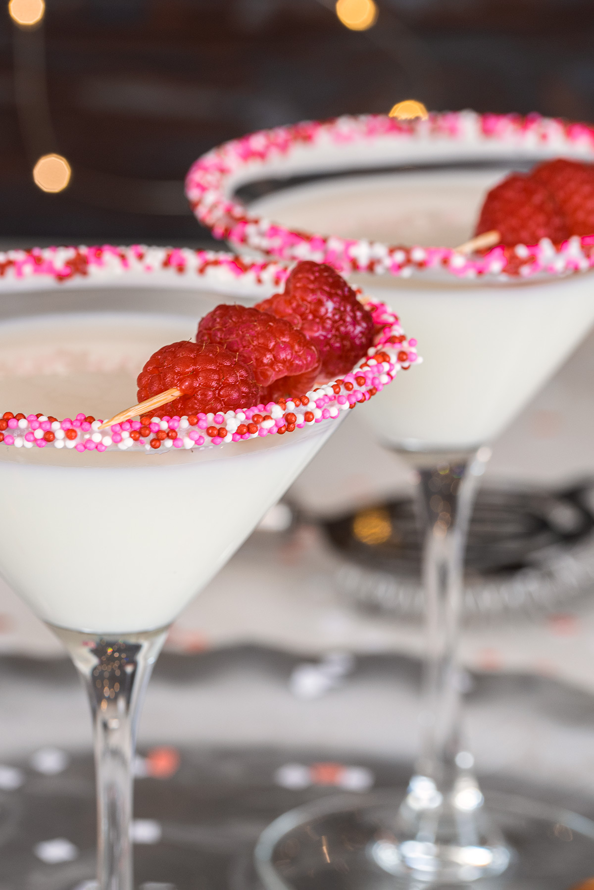 Two white chocolate martini glasses garnished with Valentine's Day sprinkles and raspberries.