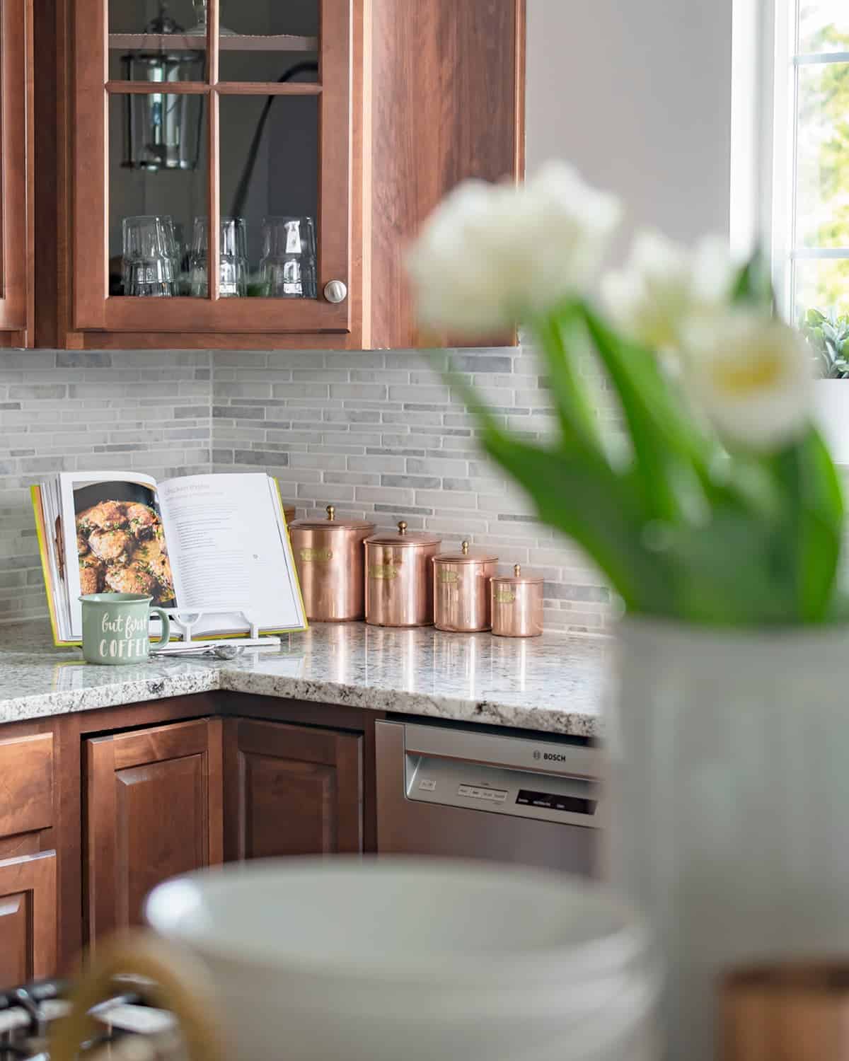 Copper canisters in modern traditional kitchen makeover on marble counter. Basket tray centerpiece on kitchen island with white bowls, copper mugs, and potted white flowers.