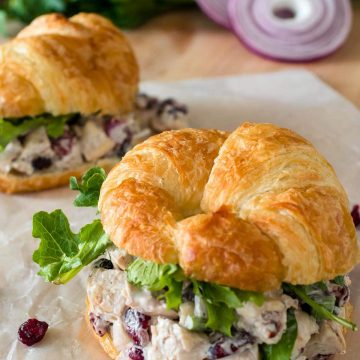 Fresh chicken salad recipe features craisins and topped with greens on croissant rolls laid on wax paper with celery and red onion in the background.
