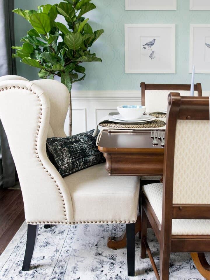 Traditional dining room reveal features plush linen wingback chair with brass tack detail and dark legs accented by modern black and white throw pillow. Traditional dining table and chairs sits on black and white floral pattern oriental rug covering dark finished floors. Light wainscotting leads up to blue-green patterned walls with framed white matted bird prints.