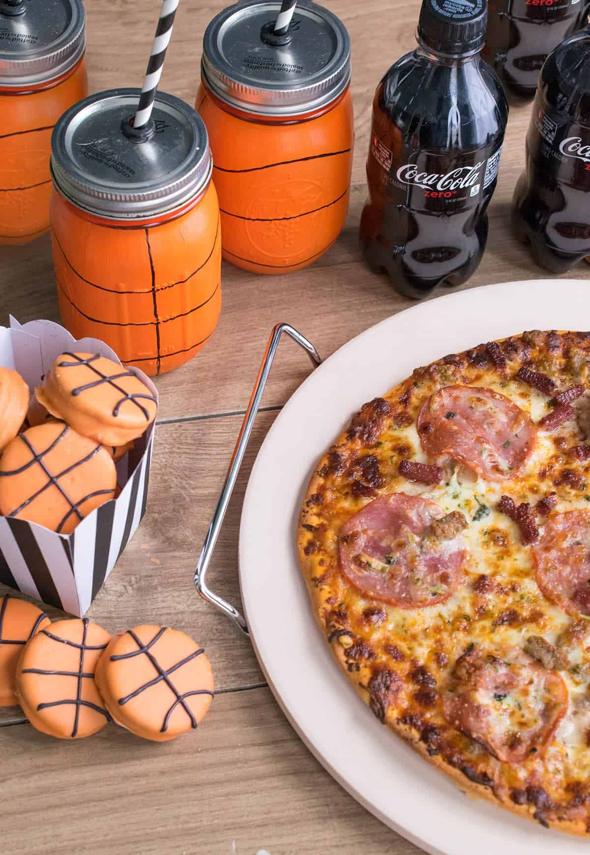 Wooden table filled with Basketball painted mason jars, basketball cookies, and bottles of Coca-cola next to pizza.