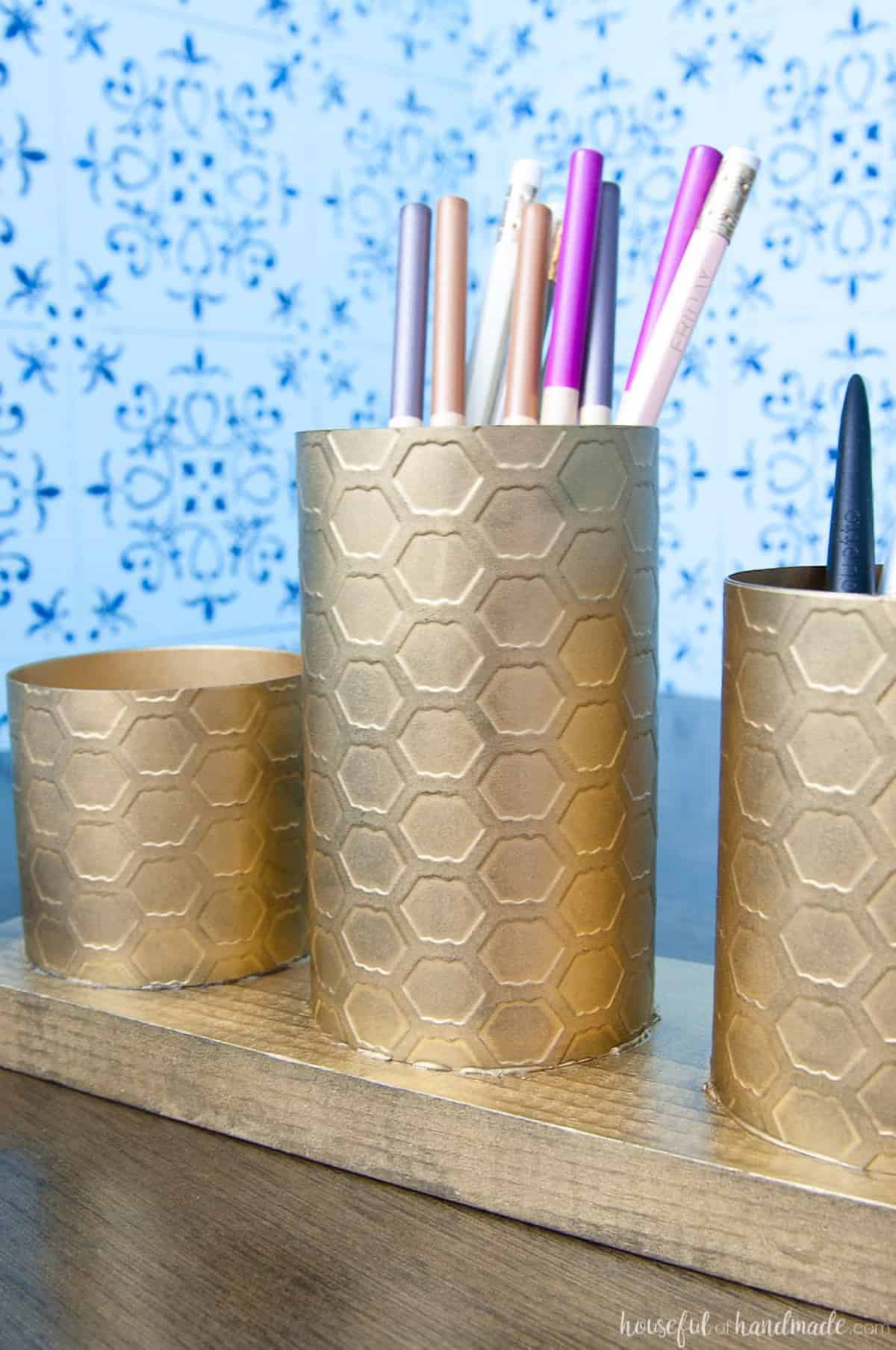 Brass painted pencil cups against blue patterned wallpaper background.