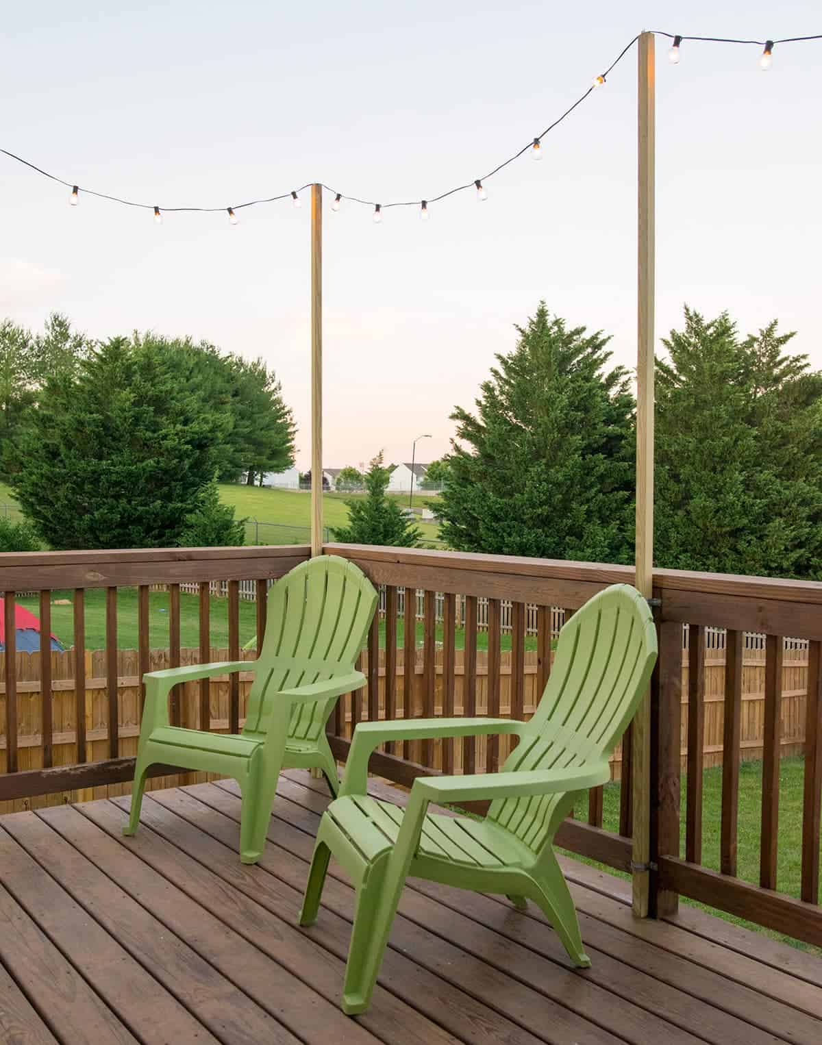 String lights hanging from posts above railing on wooden deck behind green outdoor chairs.