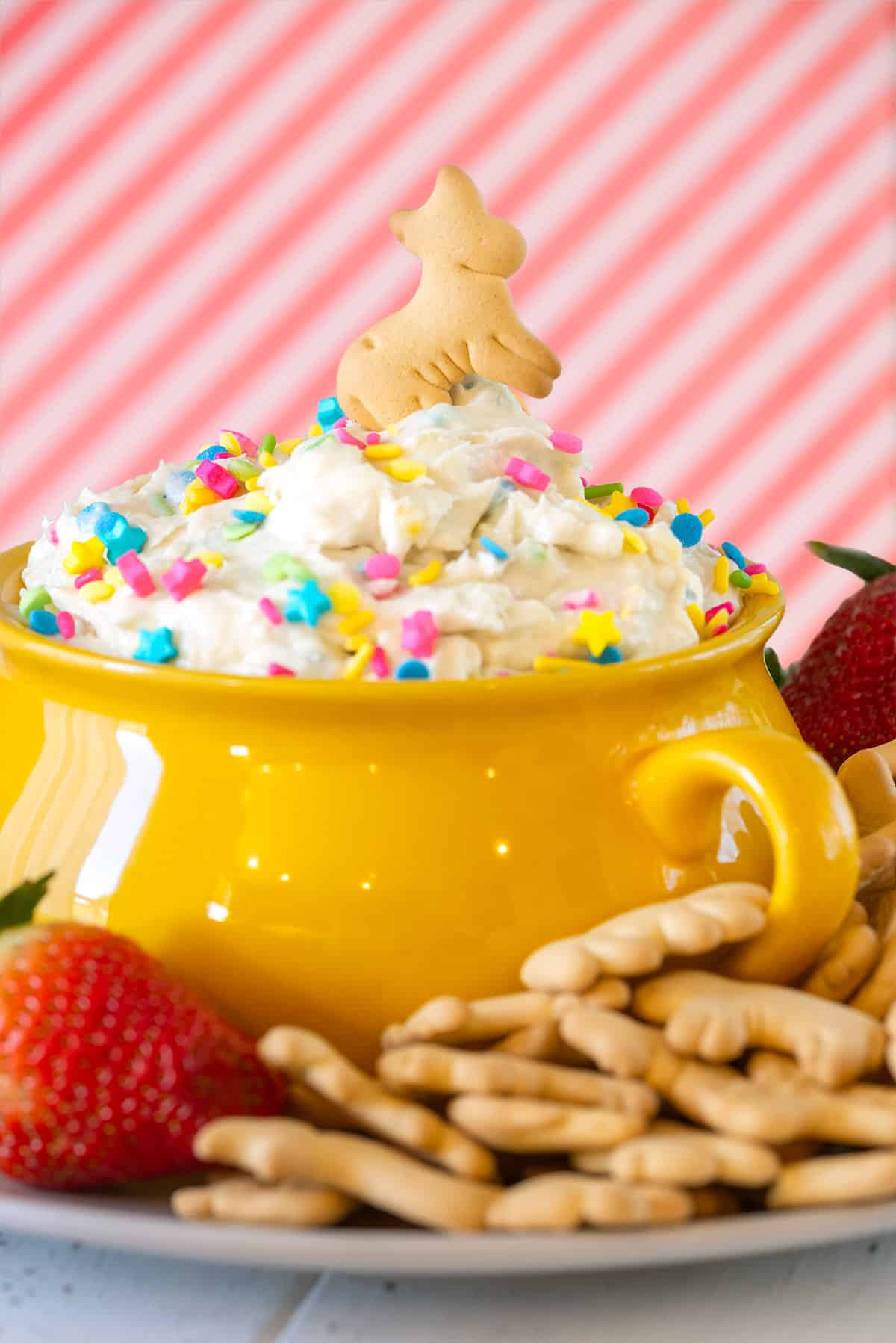 A bowl filled with funfetti dip and topped with vibrant sprinkles and a animal cracker cow. Strawberries around the side.