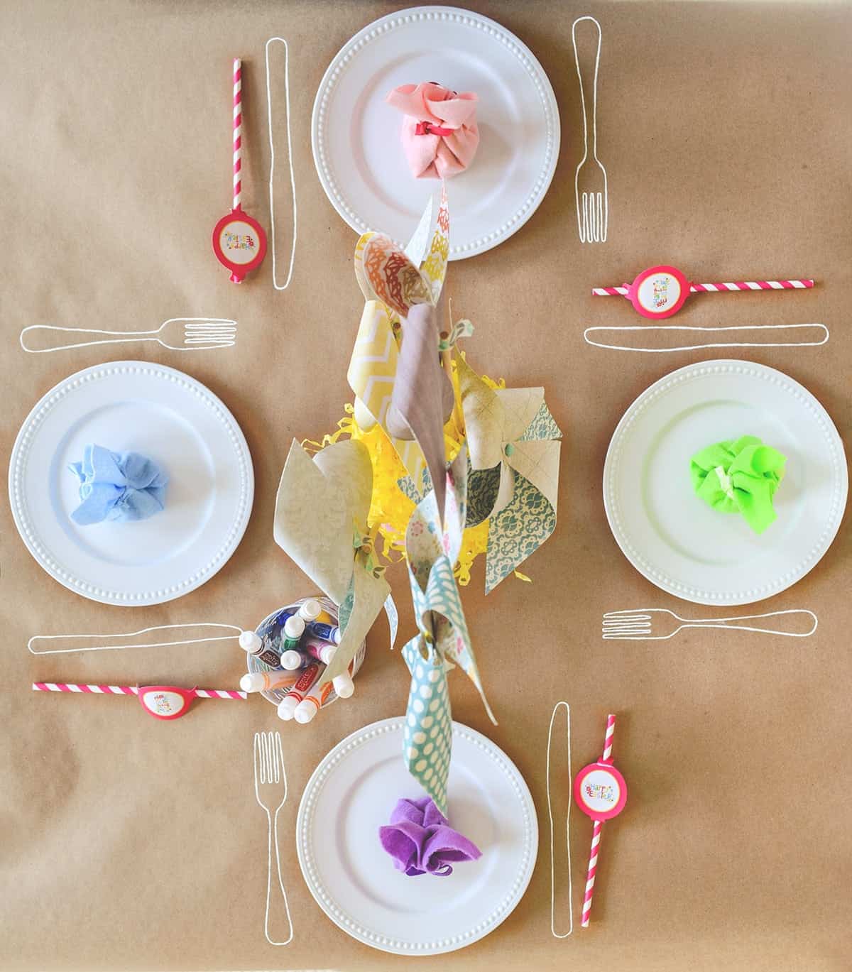 Brown craft paper table for kid's table with white plates, colorful napkins, pinwheel centerpiece, and markers for drawing on table.