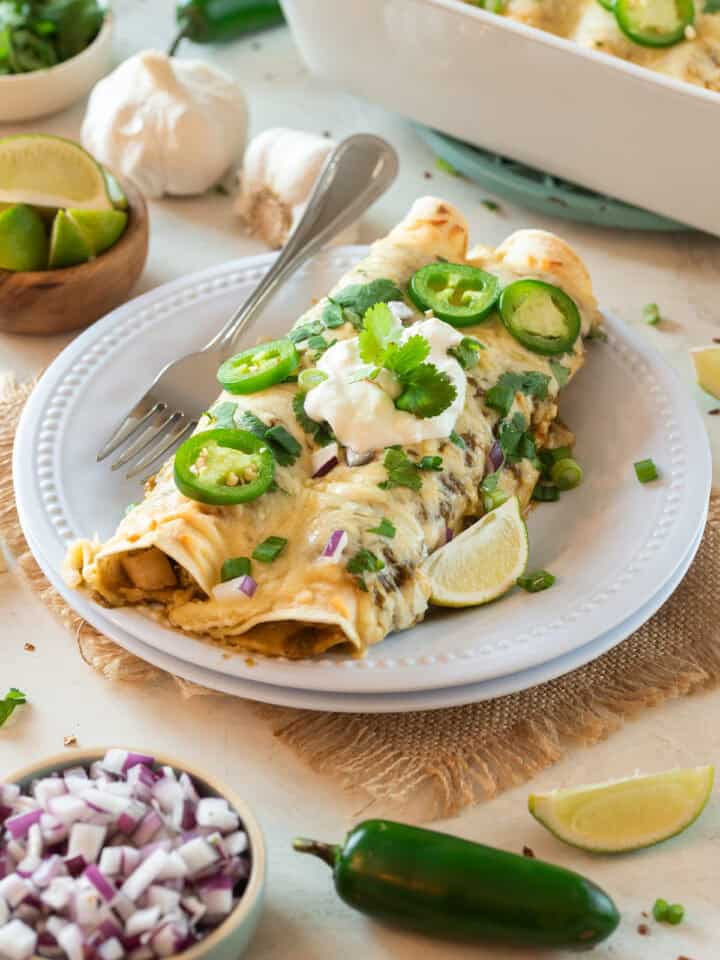 Two green enchiladas filled with chicken and topped with sour cream, cilantro, jalapeno and lime wedges on a plate.