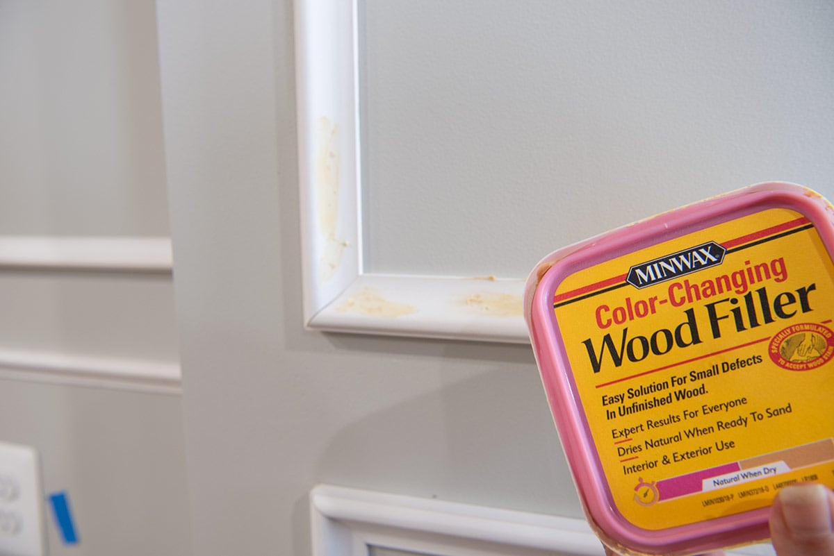 A box of wood filler held in front of small filled holes in wood trim.