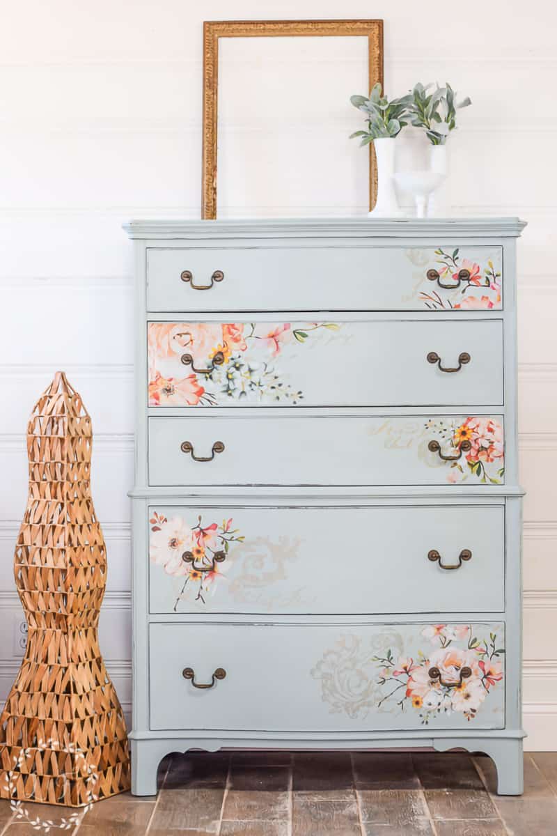Blue eggshell painted chest of drawers with pink floral accents against a white shiplap wall.