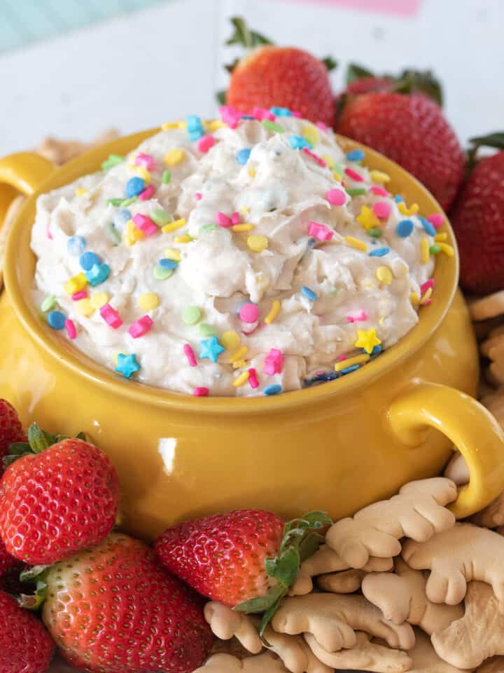 A bowl of cake batter dip with brightly colored sprinkles on top and strawberries and animal crackers around the bowl.