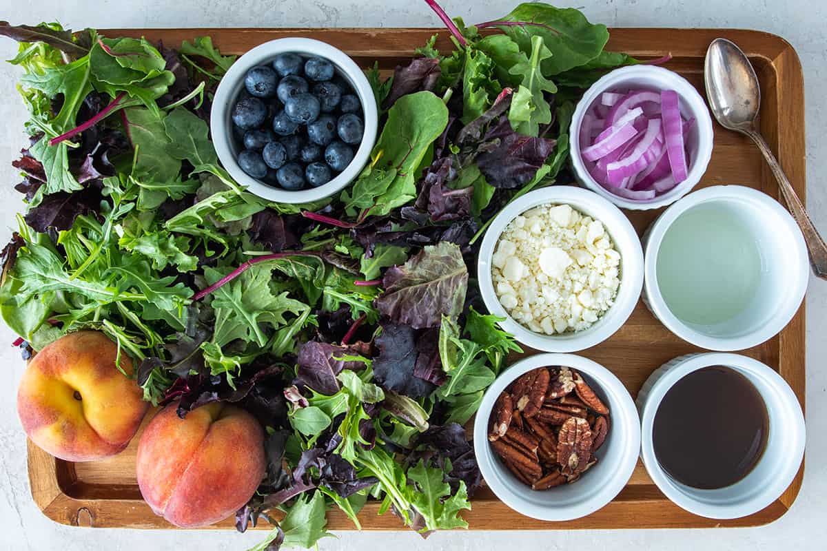 Ingredients to make spinach peach salad laid out on a board and in small bowls.