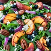 Spinach salad with grilled peaches, blueberries, cheese, pecans, and red onions on a plate.