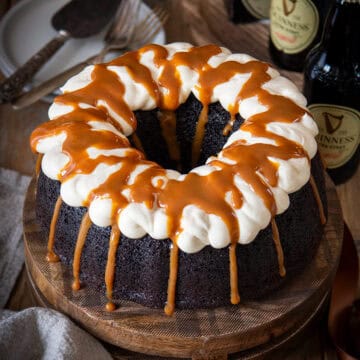 A Guinness Chocolate cake with vanilla buttercream frosting and a caramel drizzle on top in front of several Guinness bottles on a wood background.