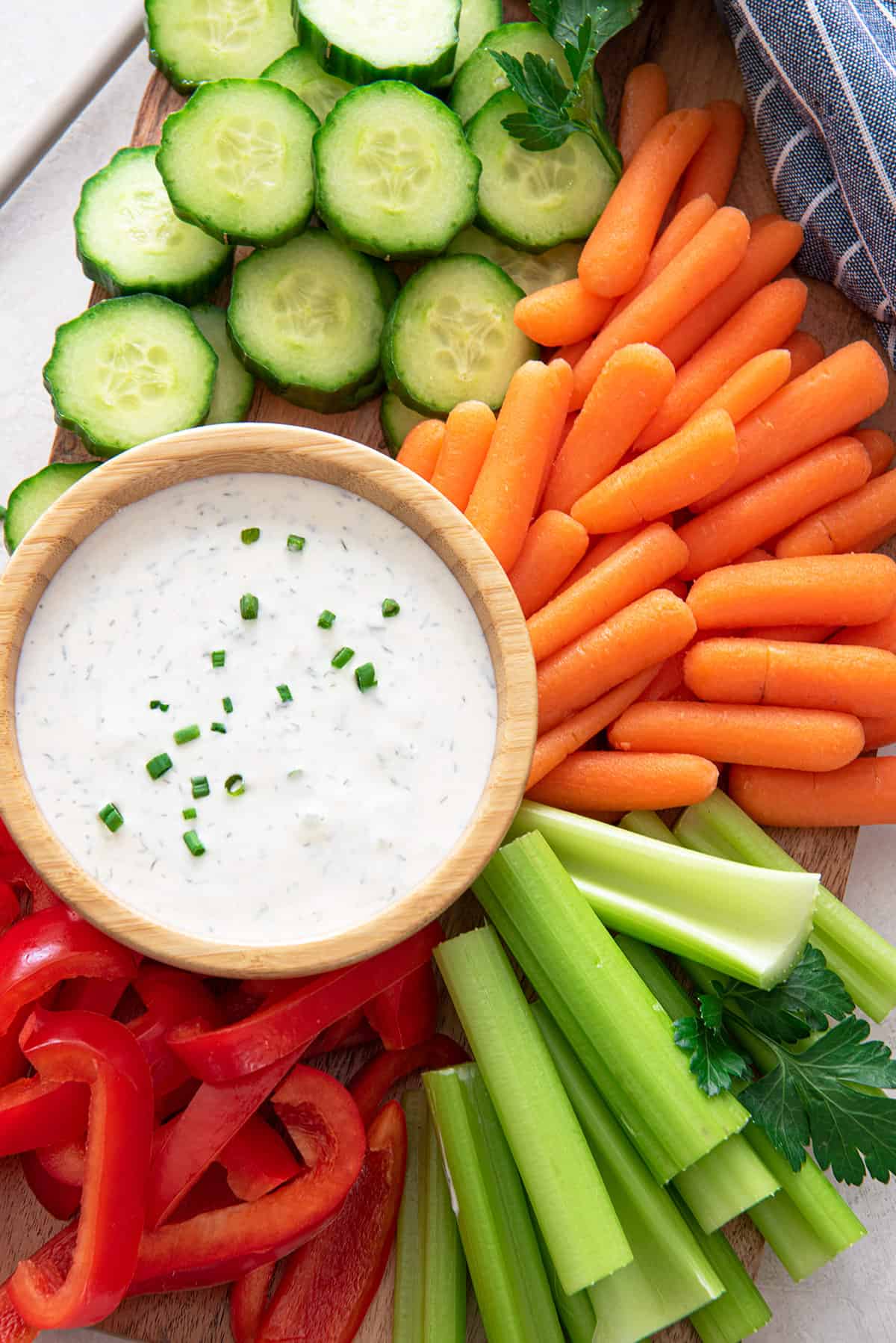 A small bowl of healthy ranch dip surrounded by cucumbers, carrots, celery, and red pepper strips on a platter.