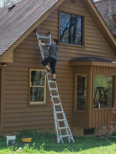 Woman on a ladder using a sprayer to stain a wood siding house exterior.