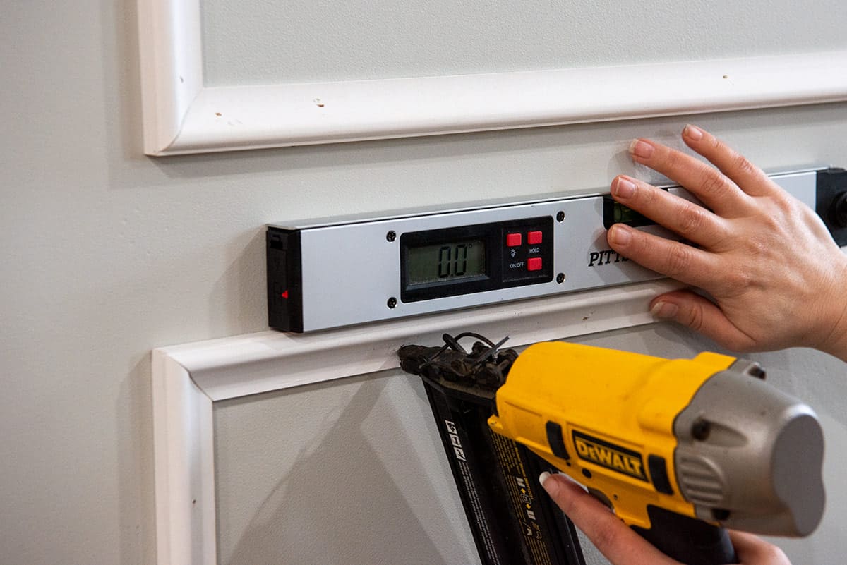 Brad nailing a picture frame molding on the wall while holding a small level.