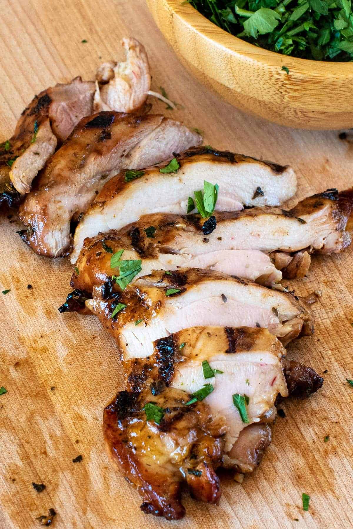 Juicy grilled and marinated chicken sliced on a wooden cutting board with a bowl of fresh herbs in the background.