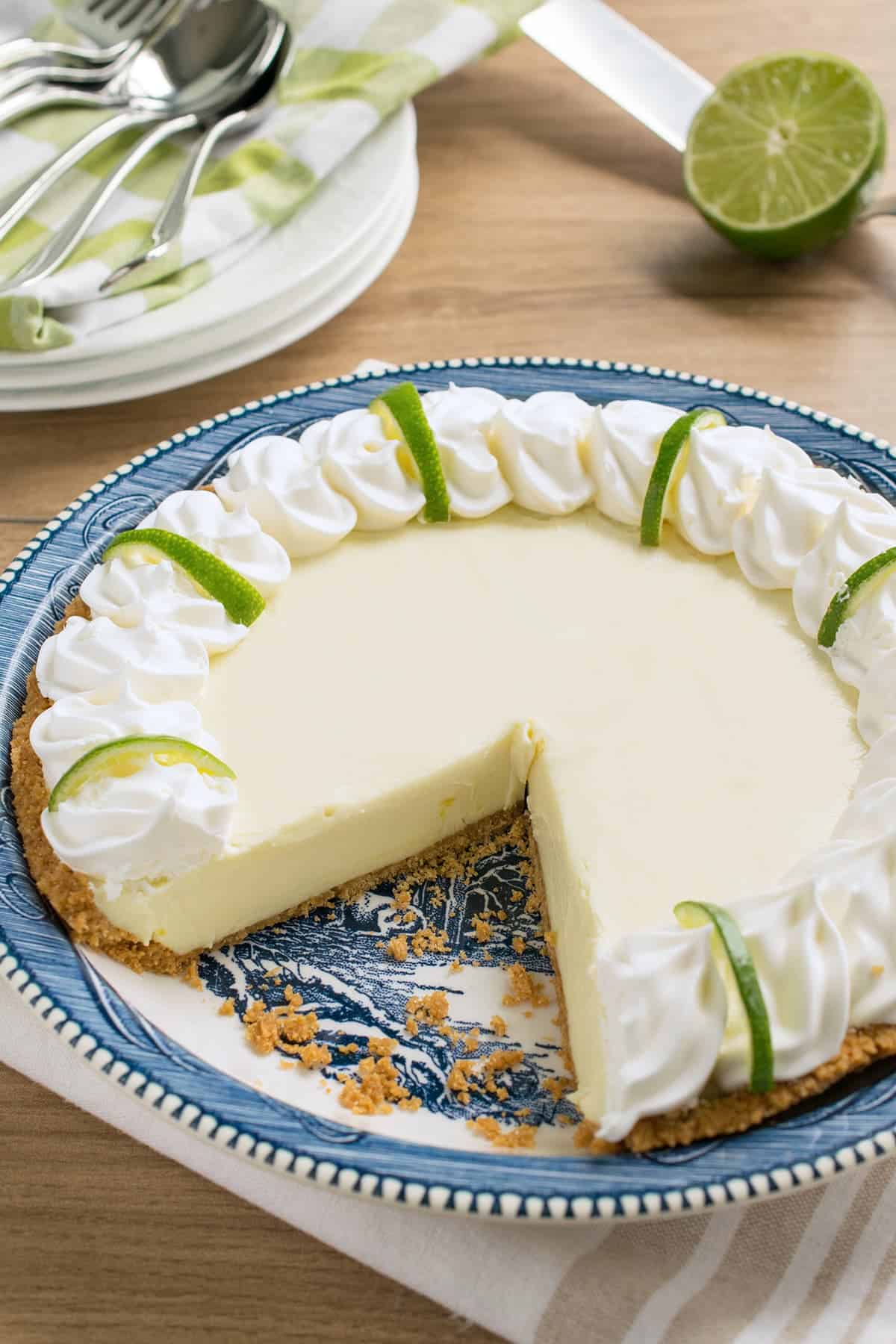 Whole lime pie in blue dish with one slice removed. Stacked white dishes with folded napkins and silverware