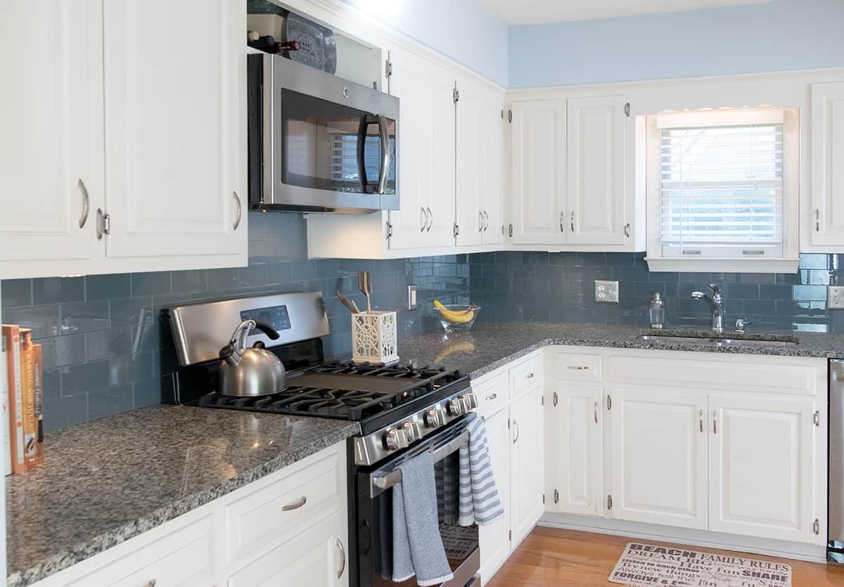Side view of full kitchen with blue glass tile for backsplash, grey countertops, and white cabinets.