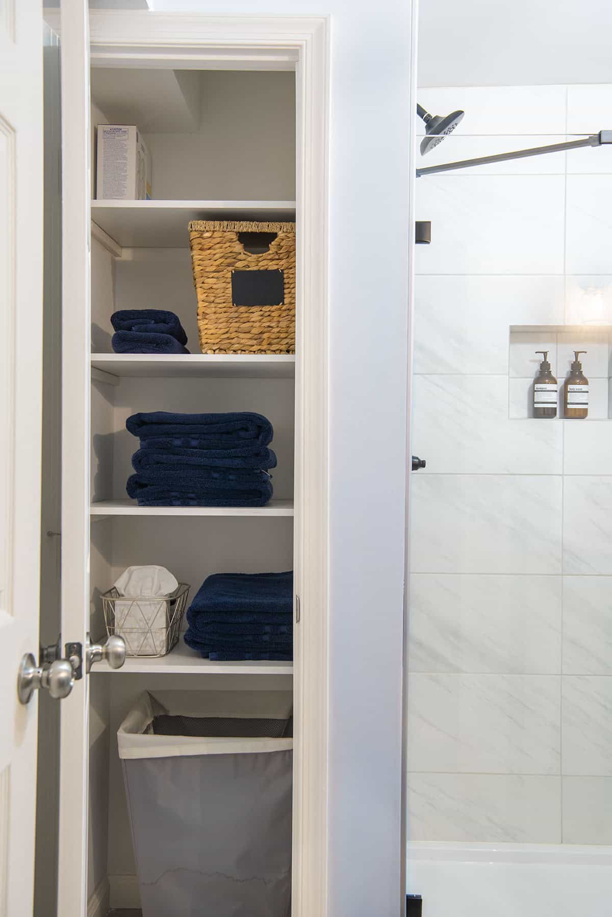A look into a modern linen closet behind a shower with blue towels, storage baskets, and a hamper for organization.