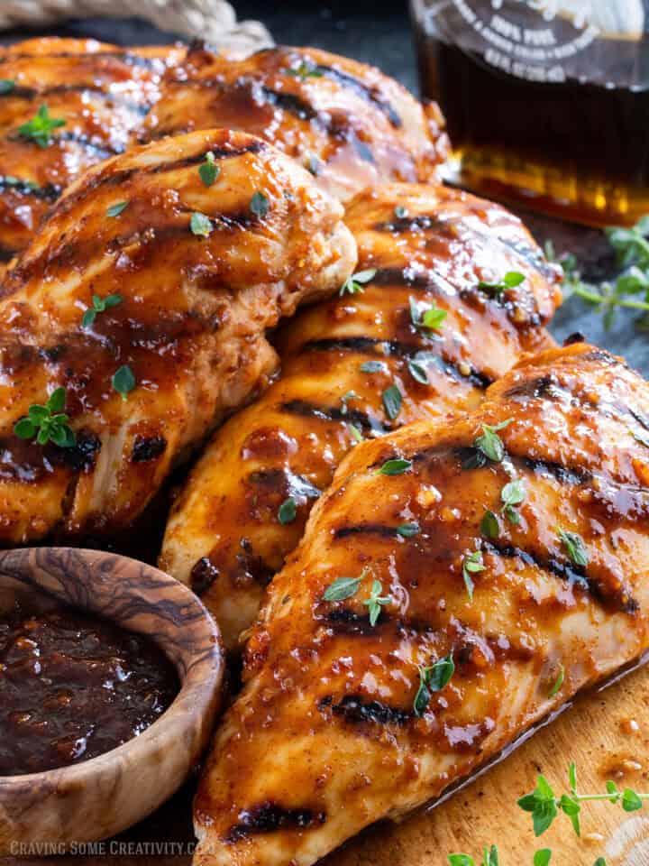 Several grilled chicken breasts with sauce on a wood board with a bottle of maple syrup.