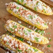 Four elotes or ears of corn dressed in mexican cheese and spices laying on a table top.