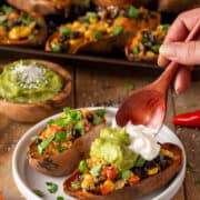 Two stuffed Mexican sweet potatoes in front while a hand tops them with guacamole and sour cream.