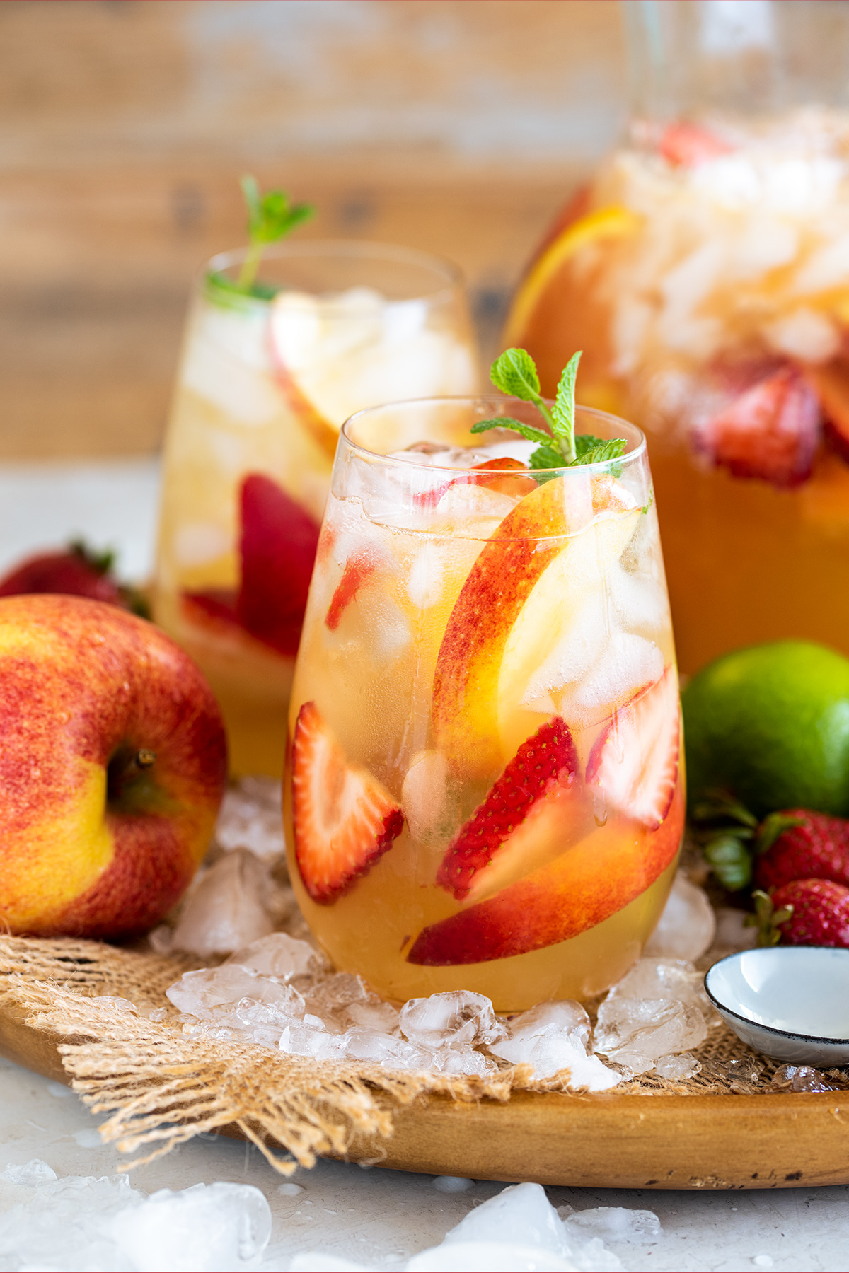 Two glasses of Moscato sangria with limes, apples, and peaches scattered around a serving tray.