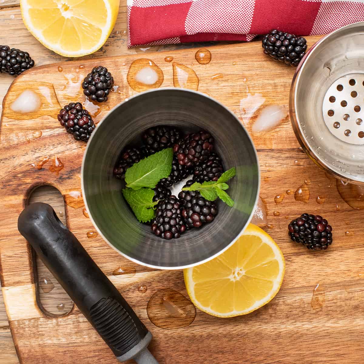 Top down view into a cocktail shaker with fruit and mint leaves in it with a cocktail muddler beside it and lemon slices around.